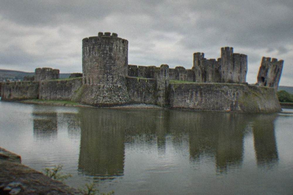 A large medieval stone castle with towers reflected in a broad water moat under an overcast sky