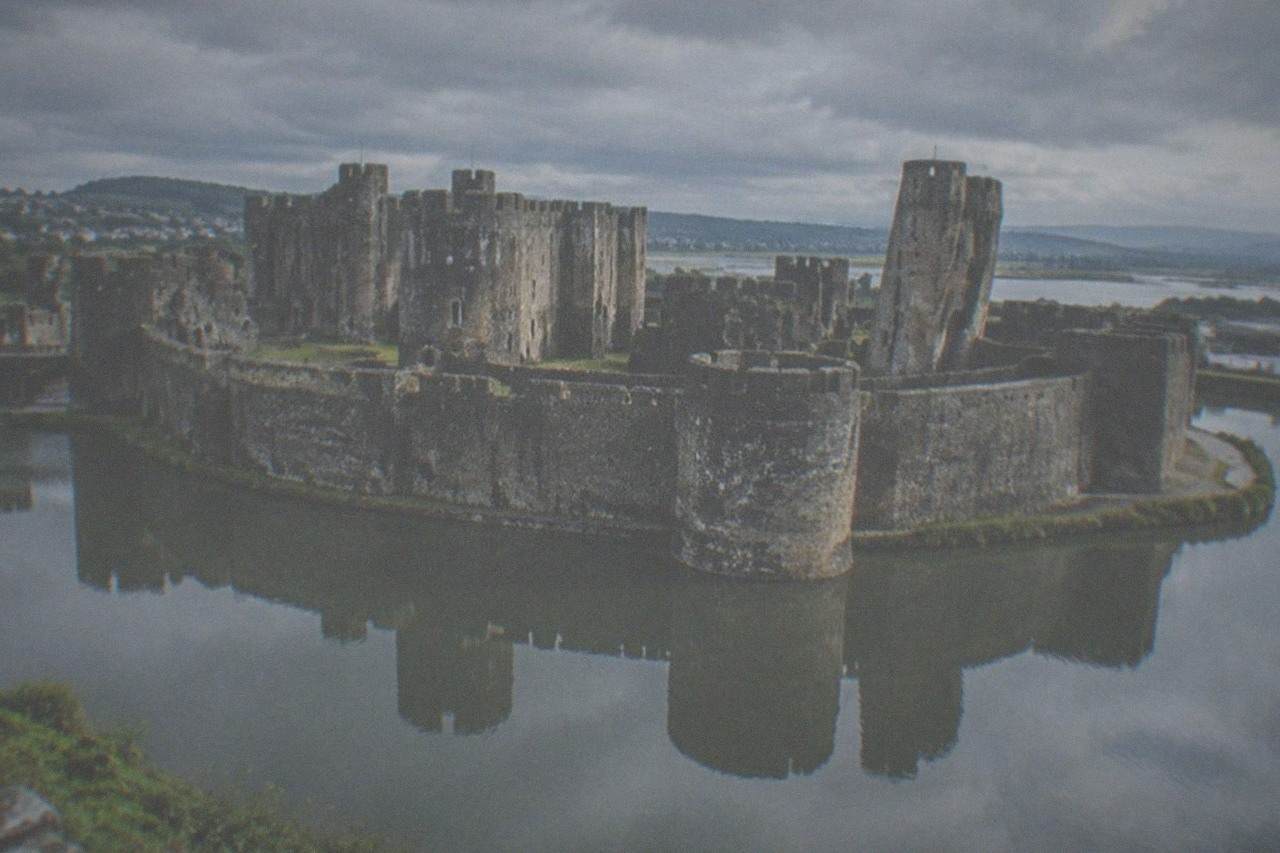 A large medieval stone castle with towers reflected in a broad water moat under an overcast sky