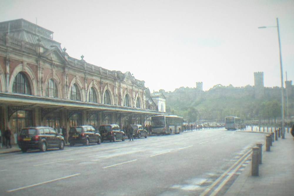 A wide city street with a stone castle wall rising behind it under a partly cloudy sky