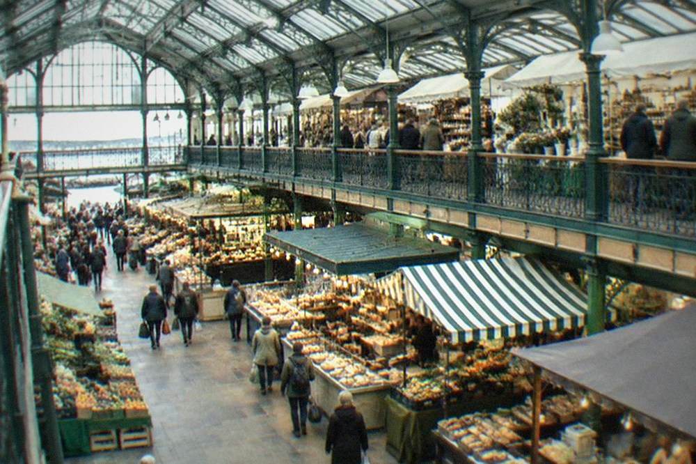 A covered Victorian market hall with food stalls and shoppers under a glass roof