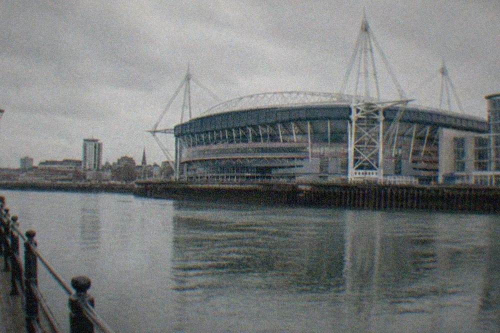 The floodlit oval roof of a large city-centre stadium rising above evening rooftops