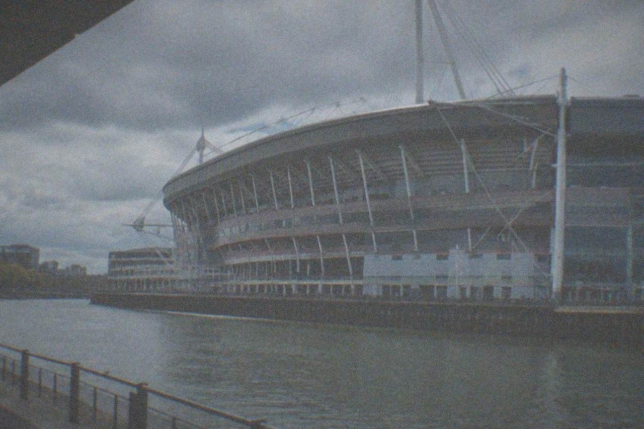 The floodlit oval roof of a large city-centre stadium rising above evening rooftops