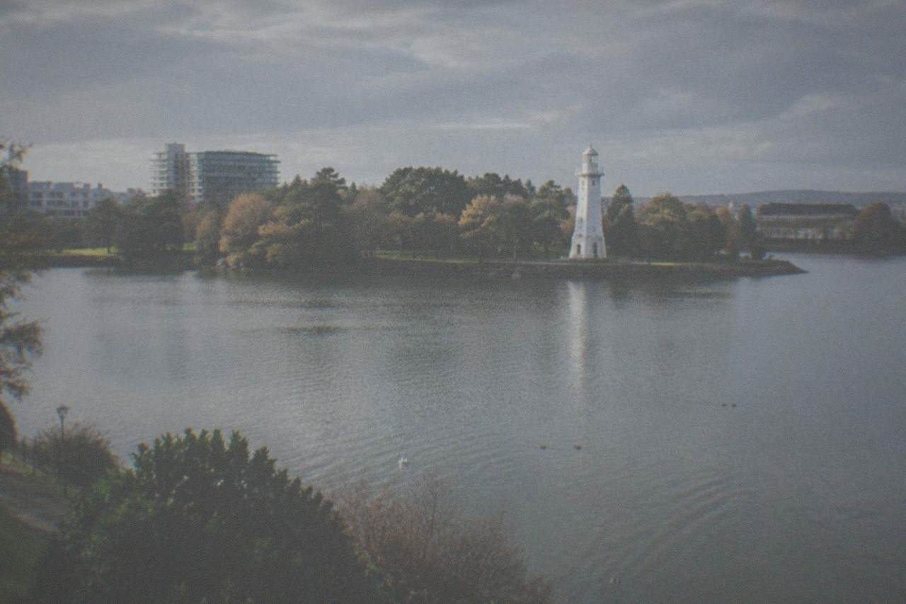 A large urban park lake with a lighthouse tower on the far shore and trees along the waterline