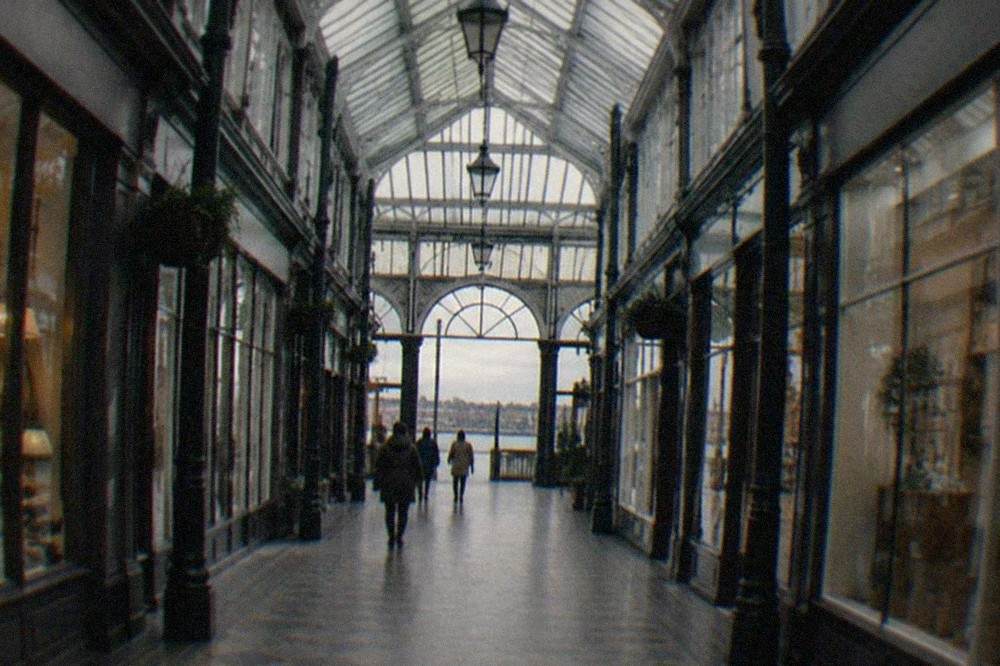 A covered shopping arcade interior with a glass roof, iron columns and small shopfronts on both sides