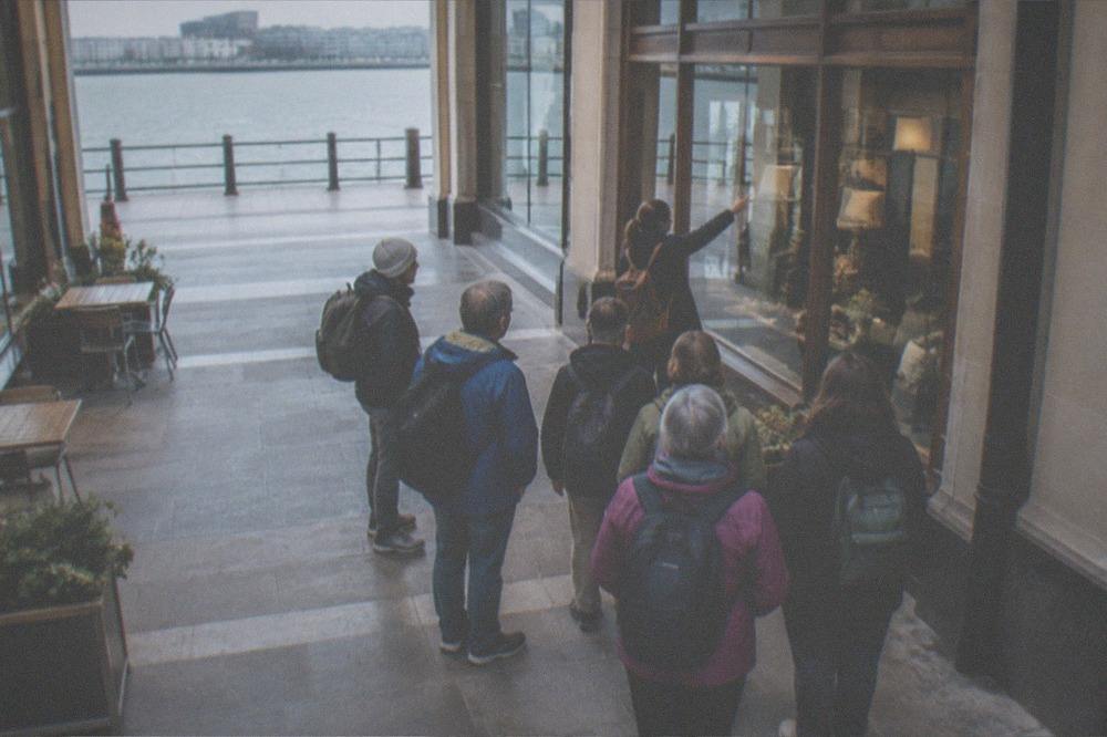 A guided walking group pausing in a covered arcade while a guide points at a shopfront
