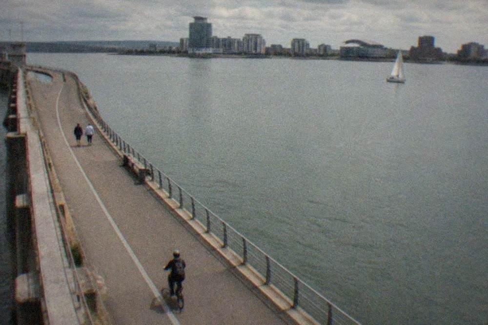 A wide waterfront path curving across a bay with a sailboat and modern buildings in the distance