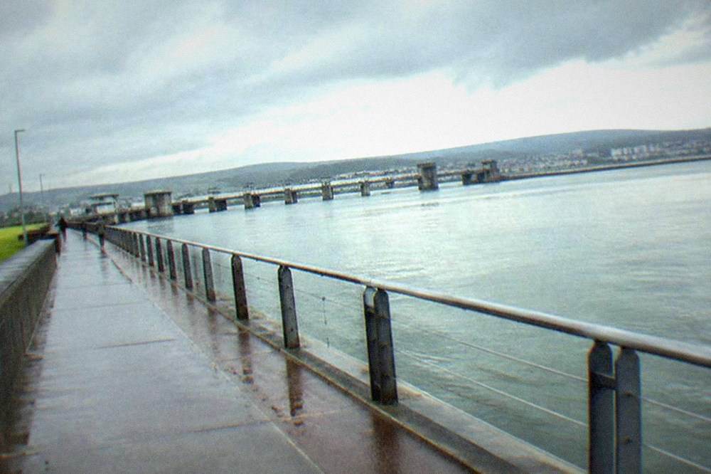 A long straight promenade beside calm water with railings and distant hills