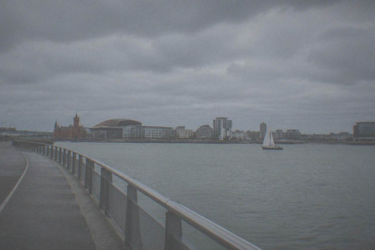 A wide waterfront path curving across a bay with a sailboat and modern buildings in the distance