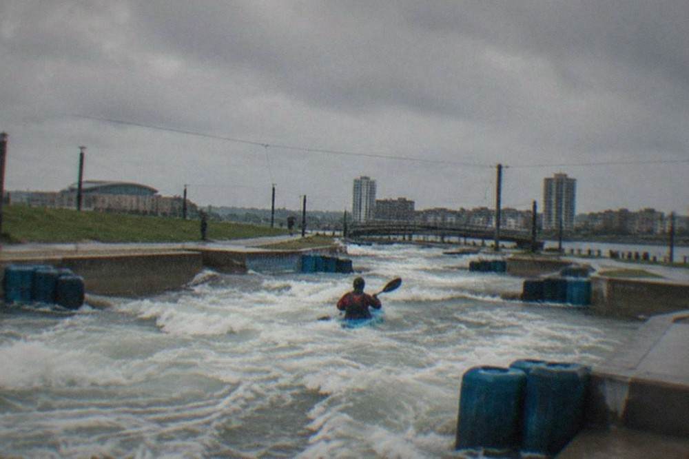 Foamy rapids rushing through a concrete white water course with grey Cardiff sky above