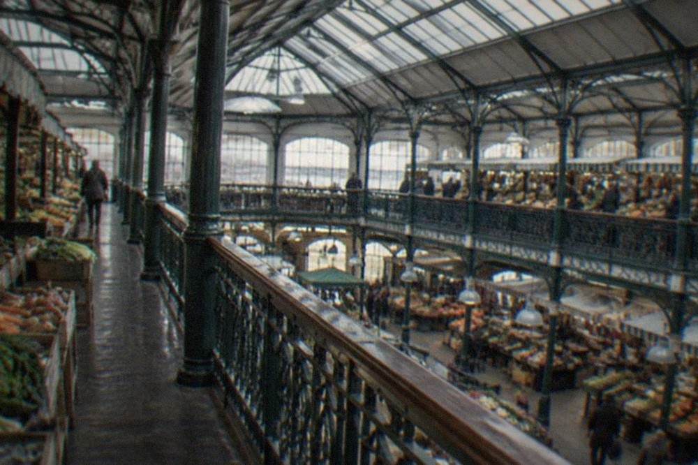 A Victorian market hall interior with a glass roof, iron columns and stalls lined with produce