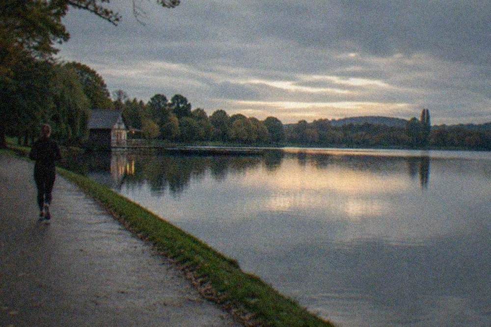 A wide park lake at dusk with trees reflected in still water and a path along the shore