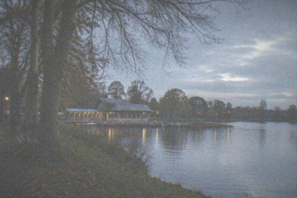 A tree lined path beside a park lake with people walking in the distance