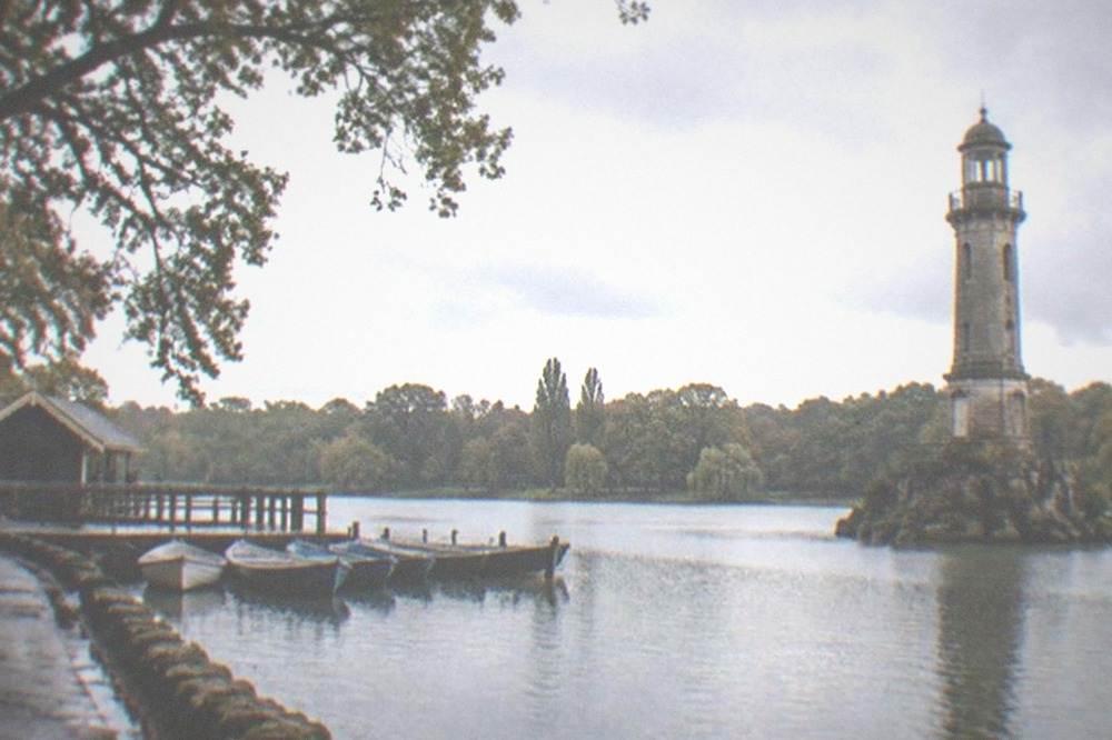 A stone lighthouse monument standing in a park lake with trees along the far shore