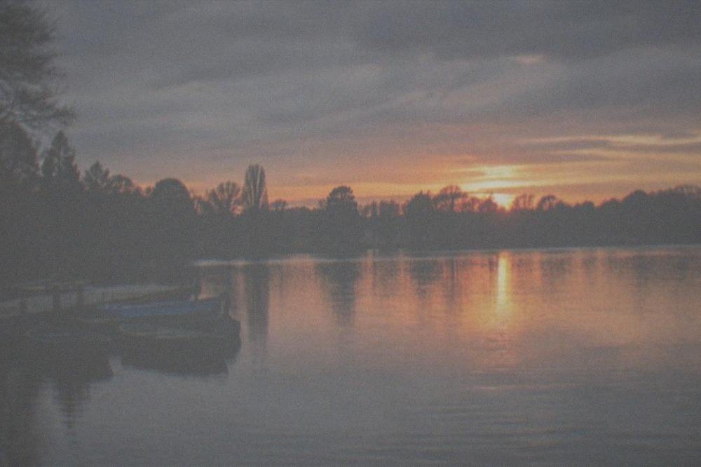 A park lake at sunset with calm water reflecting an orange sky and silhouetted trees