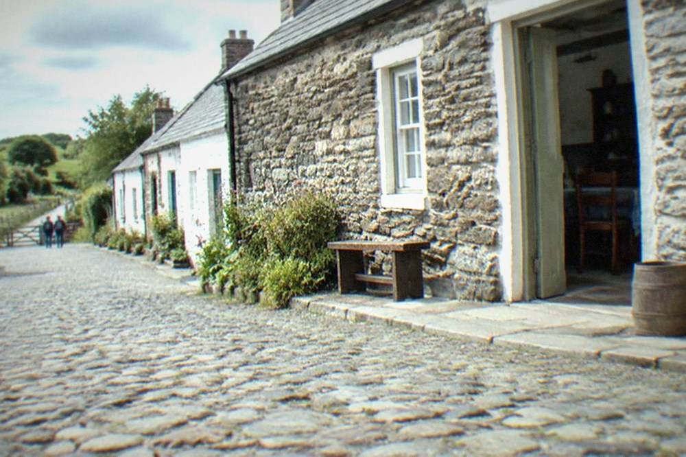 A cobbled street of small stone cottages with period furnishings visible through open doors