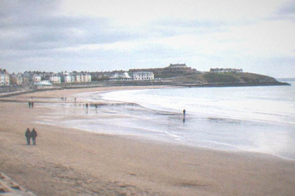 A wide sandy bay at low tide with gentle waves and a promenade in the distance