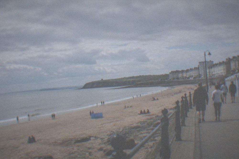 A sunny promenade with a calm sea and people walking beside beach huts