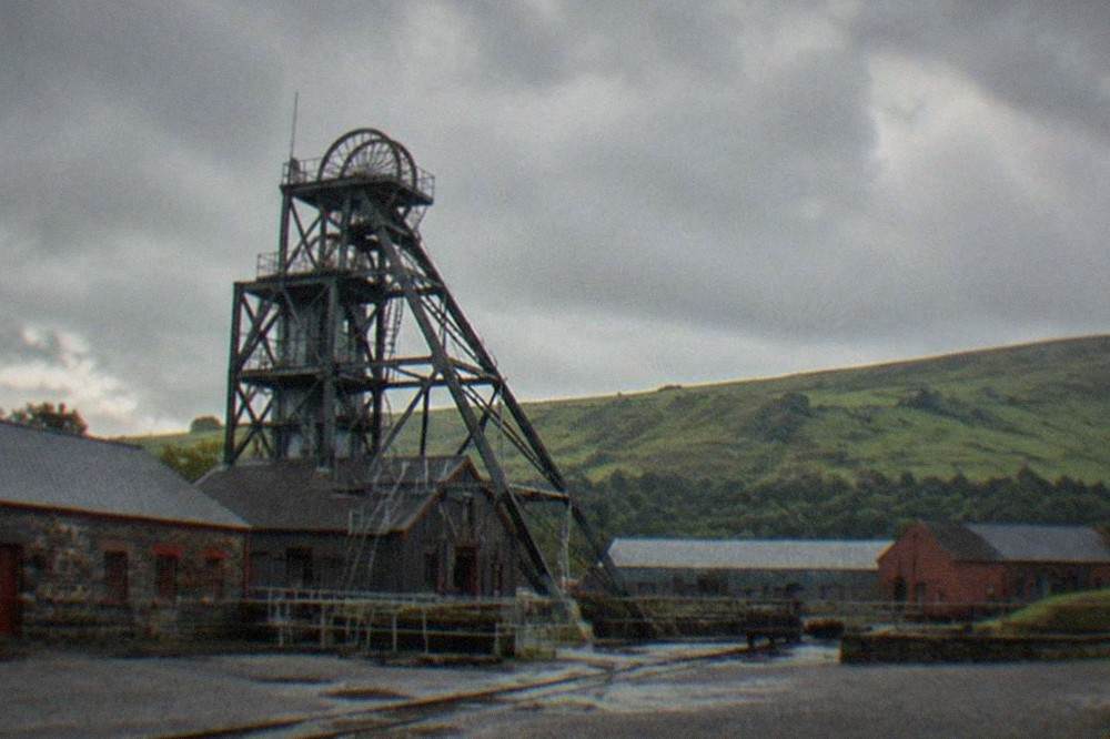 Steel winding gear tower above a colliery yard with green hills behind