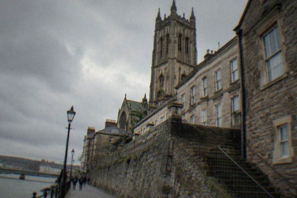 Historic stone buildings and a church tower above a riverside city street