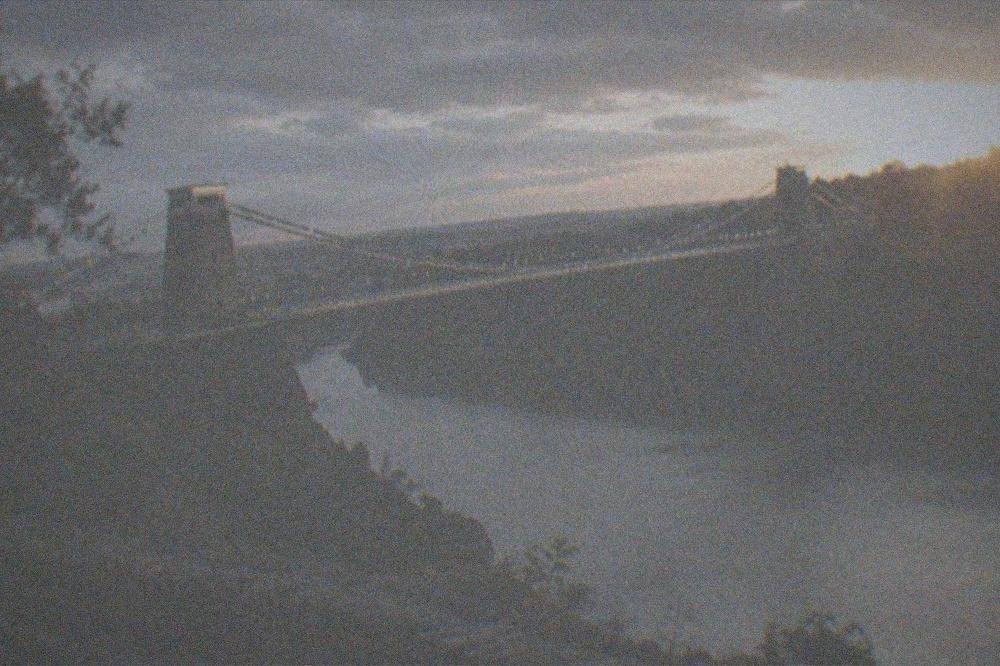 A lit suspension bridge spanning a tree-lined river gorge at dusk