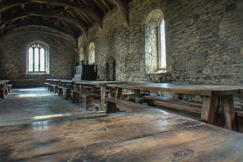 A medieval great hall interior with timber roof, stone walls and long empty tables