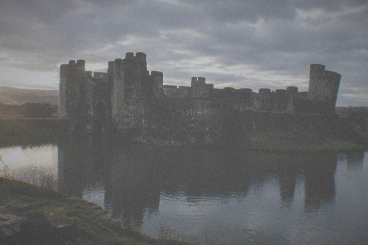 A vast medieval castle with twin towered gatehouses and a wide water filled moat under a pale sky