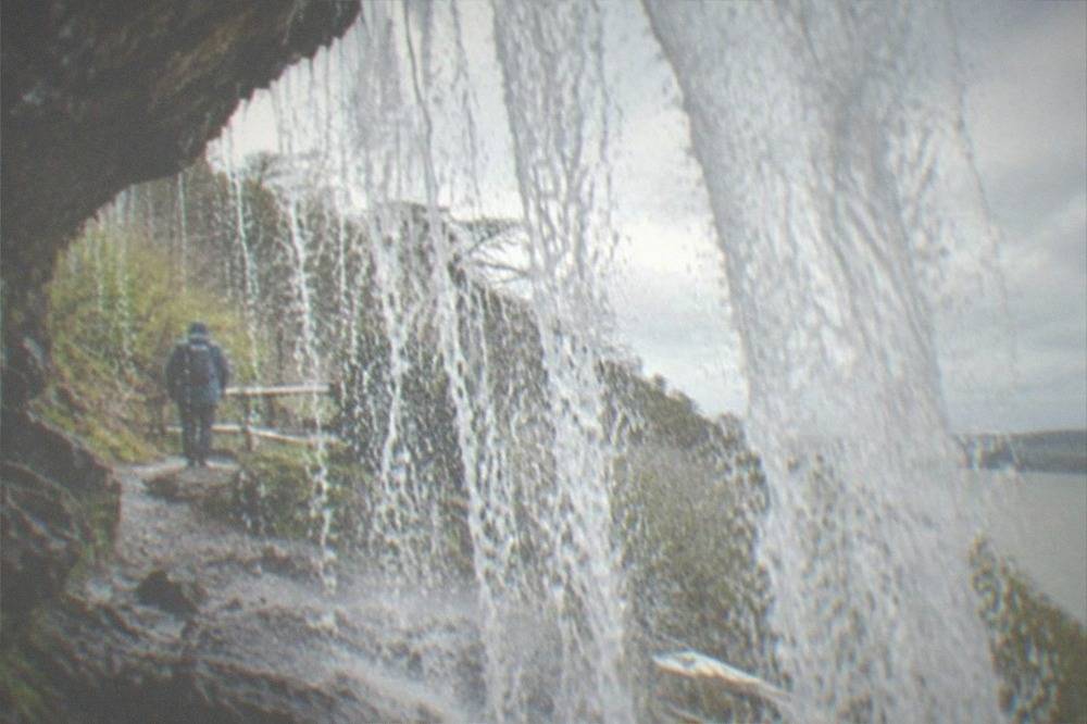 A walker standing on the path behind a curtain of falling water at a waterfall