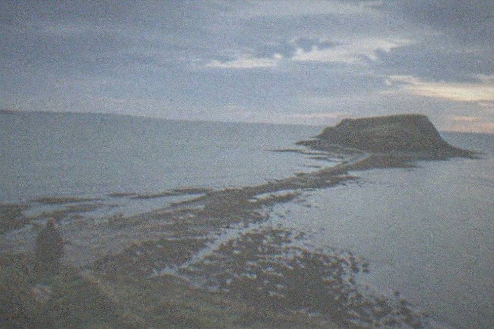 A flat rocky causeway stretching from a grassy headland towards a small island