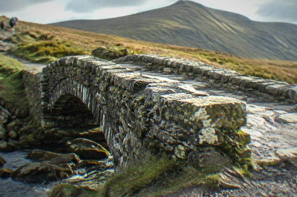 A stone footbridge crossing a stream at the base of a mountain path