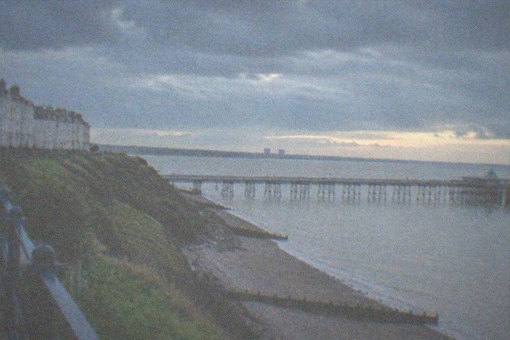 A shingle beach below grassy cliffs with a pier in the distance