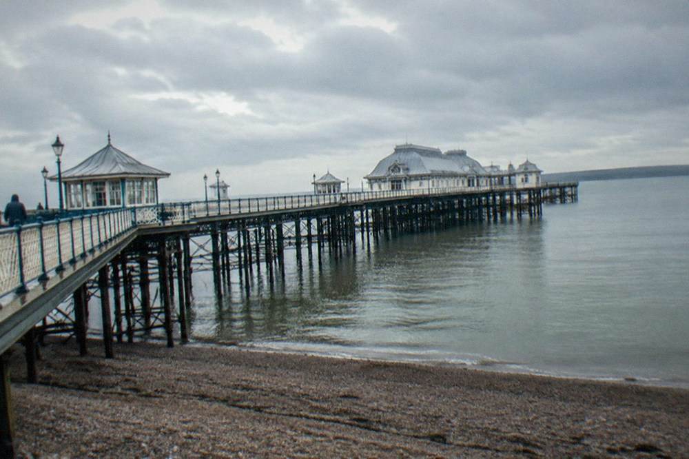 A Victorian pier extending over a shingle beach on a calm overcast morning