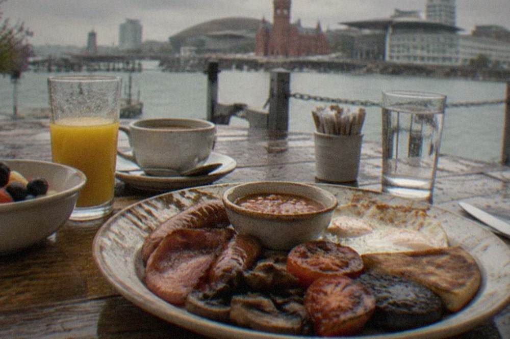 A wooden table set for brunch with glasses, fruit and a full Welsh plate