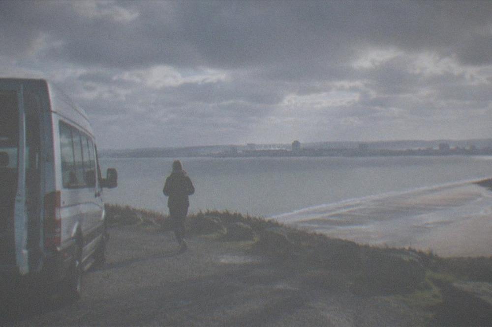 A tour minibus parked at a coastal viewpoint above a wide sandy bay in South Wales