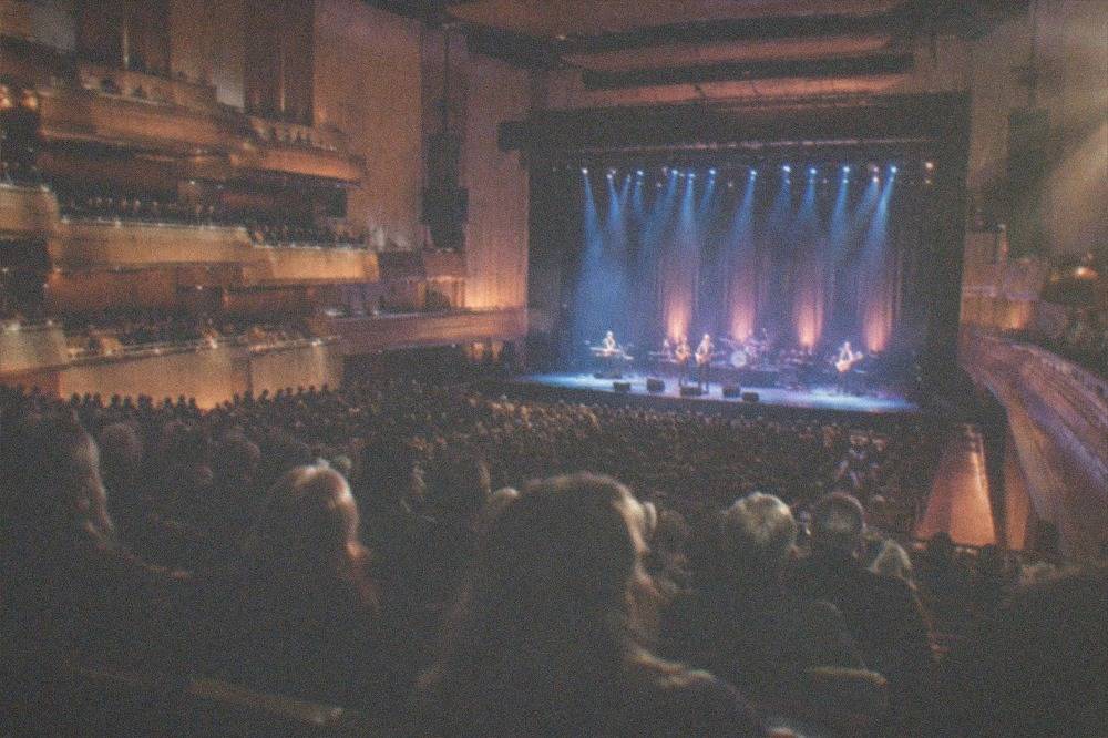 A concert crowd facing a lit stage inside a large hall
