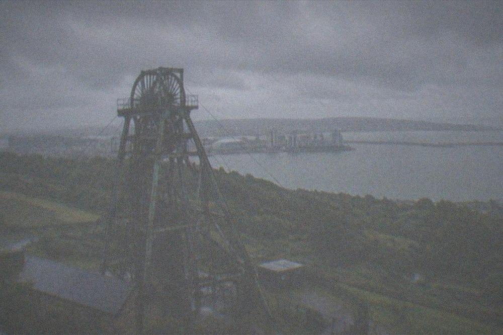 A metal mine headframe tower standing over green hills under a wide sky