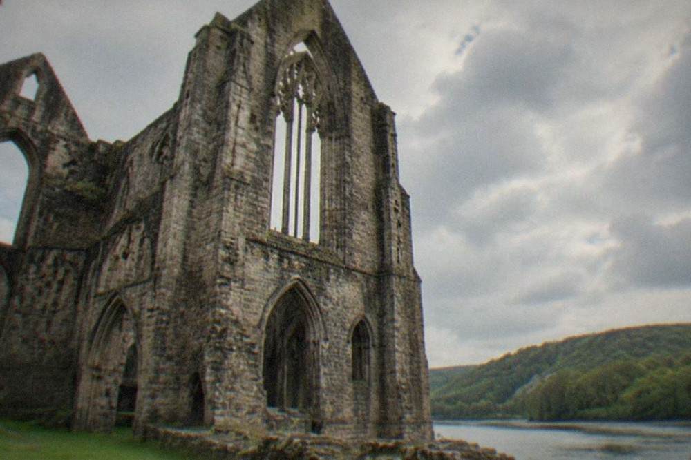 A ruined gothic abbey beside a river with forested hills behind it