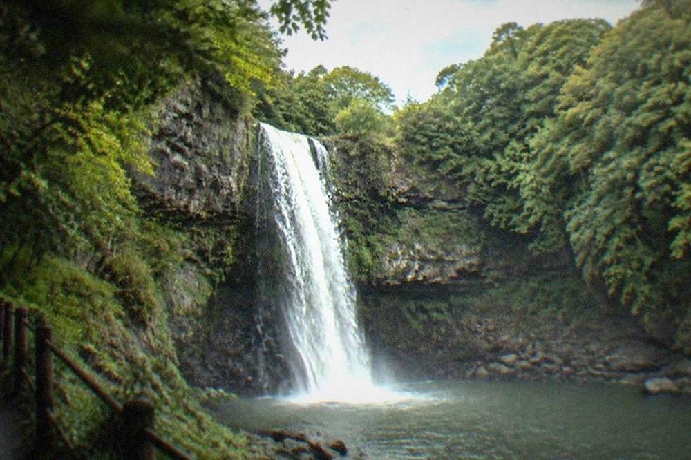 A tall waterfall dropping into a rocky wooded gorge with a path beside it