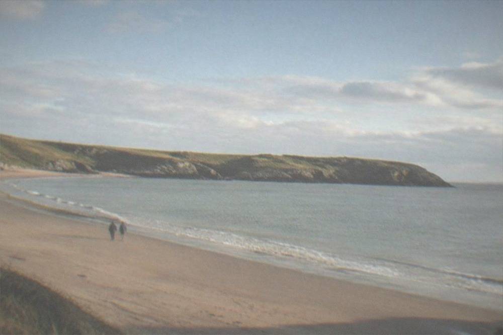 A wide sandy beach curving between grassy headlands under a clear sky