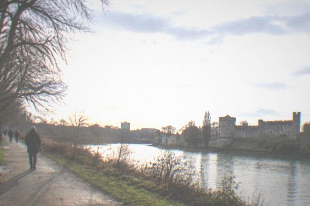 A tree-lined park path beside a river with a castle wall in the distance