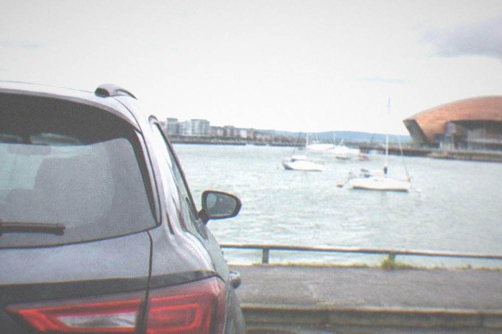 Waterfront car park beside a wide bay with sailboats and a modern building with a bronze facade