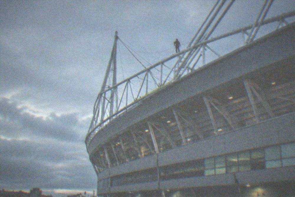 A person in a harness walking along a high gantry above a stadium roof