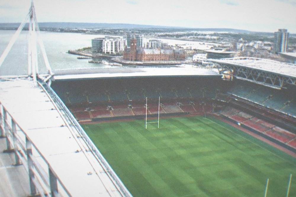 A rugby stadium pitch and stands seen from a high rooftop walkway above the roof