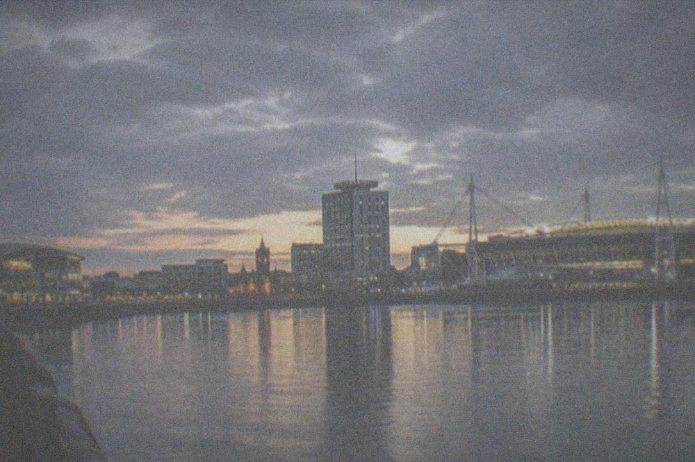 A city skyline at dusk with a stadium roof lit up beside a river