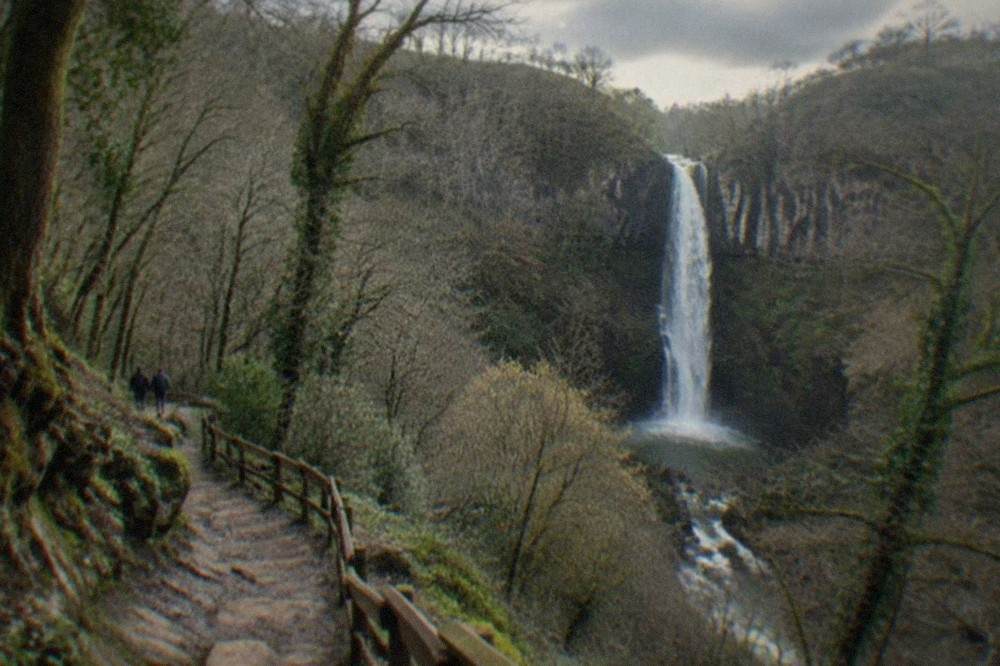 A forest path leading towards a tall waterfall in a wooded valley