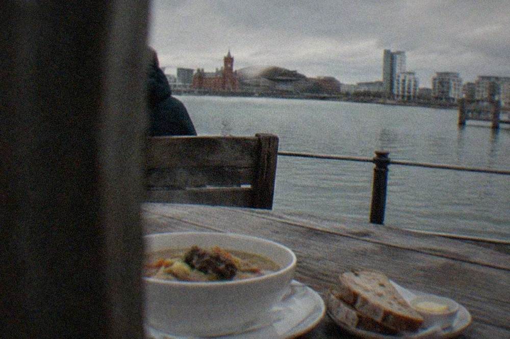 A bowl of steaming lamb cawl beside sliced bread and butter on a wooden pub table