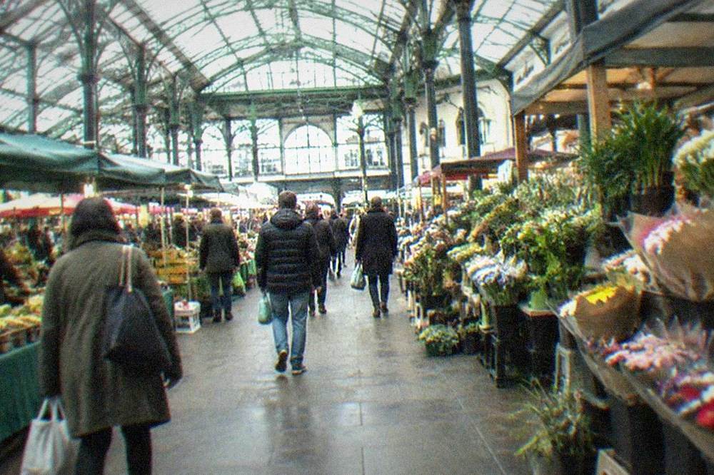 A Victorian market hall interior with stalls beneath a glass roof and shoppers carrying bags