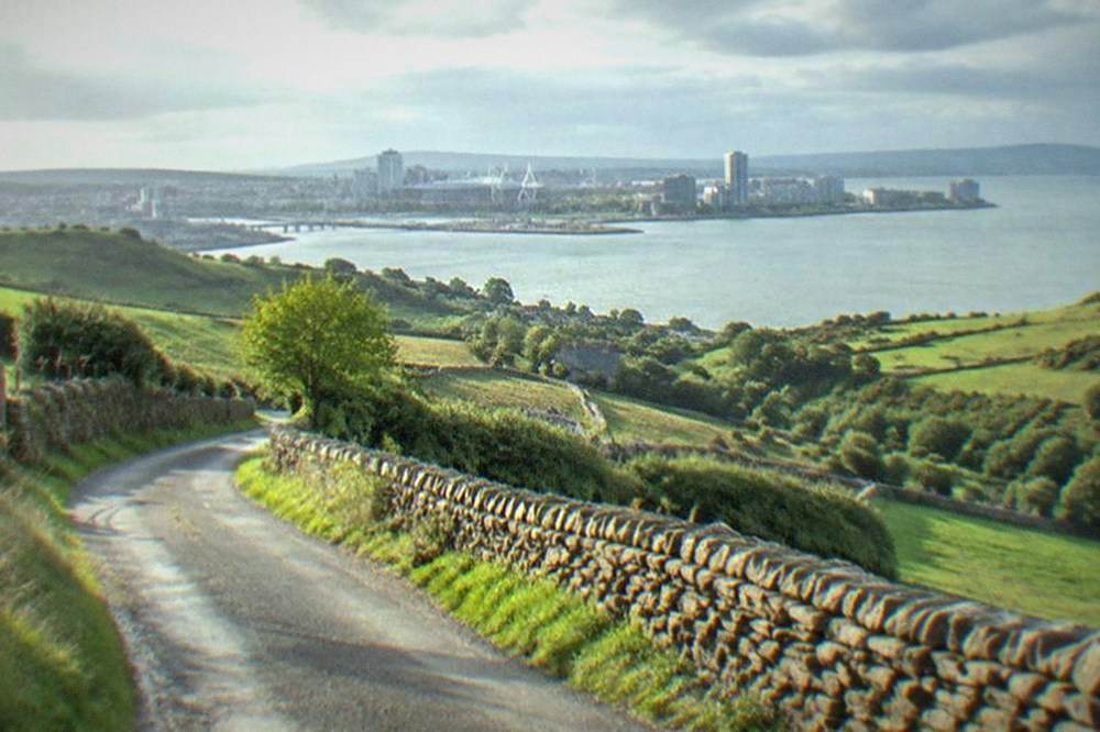 A narrow country lane lined with dry stone walls curving between green hills